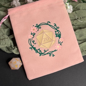 Puede incluir: Una bolsa de color rosa claro con cordón que presenta un gráfico central de un dado de veinte caras dorado encerrado en una corona de enredadera verde oscuro adornada con pequeñas flores rosas. Un dado de veinte caras blanco translúcido está al lado.