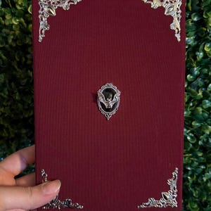 Puede incluir: Un libro de color burdeos con adornos de esquina plateados y un emblema de murciélago. La cubierta tiene una apariencia texturizada. El emblema del murciélago está centrado en la cubierta y está rodeado por un marco plateado decorativo.