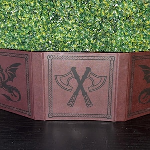 May include: A burgundy, tri-fold tabletop game screen. The screen features black dragon and axe graphics. The corners are reinforced with gold-colored metal. The background is a green hedge.