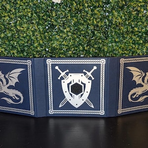 May include: A navy blue, three-panel tabletop screen with silver accents. The panels feature a dragon, shield, and sword design, suitable for tabletop role-playing games. The screen is set against a backdrop of green foliage.