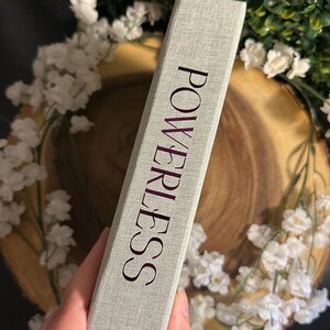 Op de afbeelding: Een grijs boek met een linnen omslag met het woord "POWERLESS" in roze op de rug.