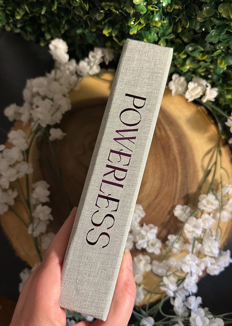 Op de afbeelding: Een grijs boek met een linnen omslag met het woord "POWERLESS" in roze op de rug.