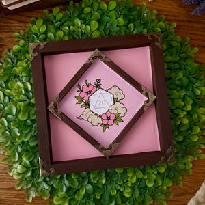 May include: A square wooden frame with dark brown edges and ornate metal corner accents. Inside, a diamond-shaped inner frame surrounds a pink background featuring a central illustration of a twenty-sided die surrounded by pink flowers and clouds.