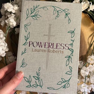 Op de afbeelding: Een grijs hardback boek met een zilveren zwaard en groene bladerenranken op de omslag. De titel "POWERLESS" is in paars gedrukt en de auteur "Lauren Roberts" in zwart.