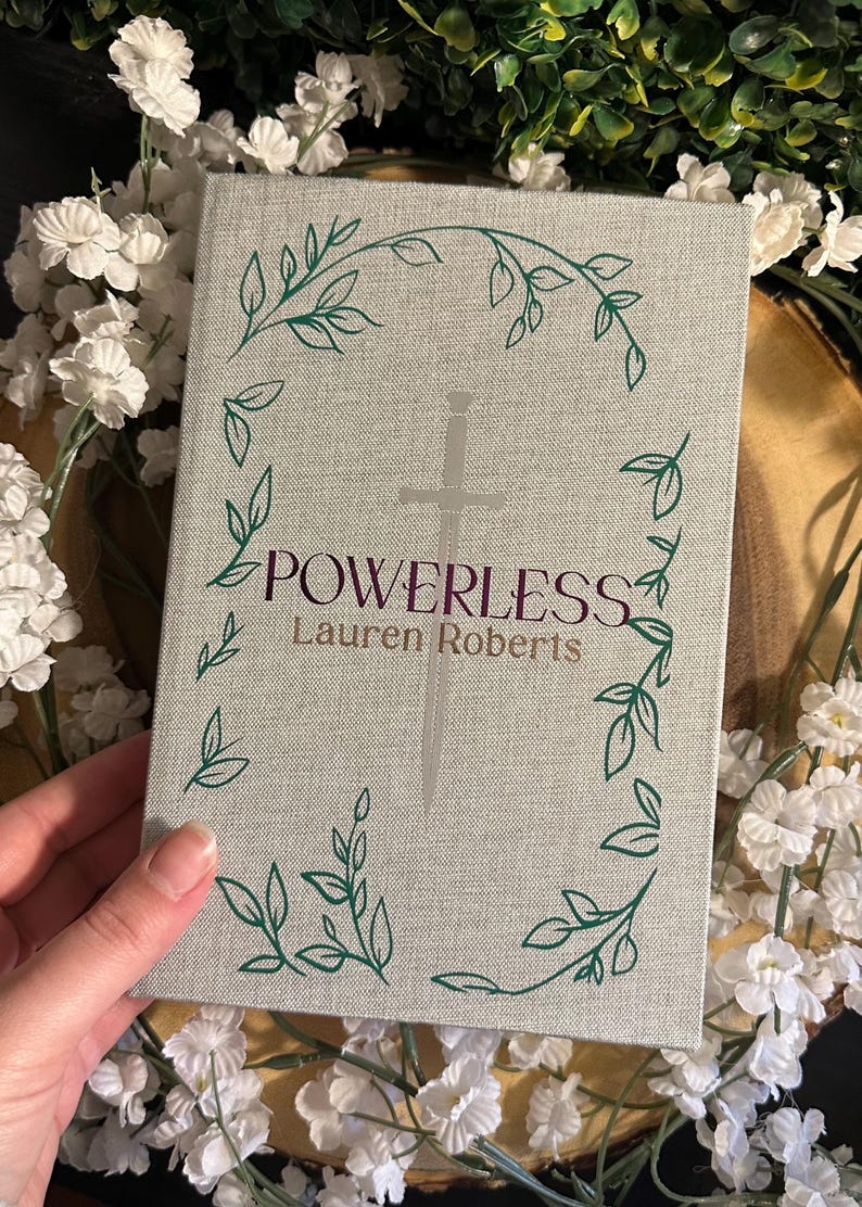 Op de afbeelding: Een grijs hardback boek met een zilveren zwaard en groene bladerenranken op de omslag. De titel "POWERLESS" is in paars gedrukt en de auteur "Lauren Roberts" in zwart.