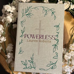 Op de afbeelding: Een boek getiteld "POWERLESS" van Lauren Roberts. De omslag is lichtgrijs met een zilveren zwaard en groene ranken.