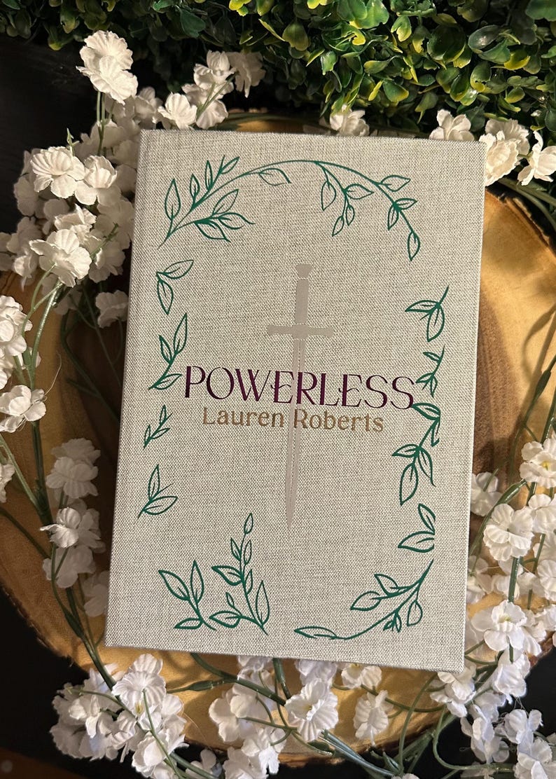 Op de afbeelding: Een boek getiteld "POWERLESS" van Lauren Roberts. De omslag is lichtgrijs met een zilveren zwaard en groene ranken.
