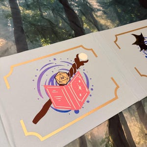 May include: A light blue folding screen panel featuring a gold-bordered rectangular frame. Inside the frame is a graphic illustration of a brown wooden staff topped with a white orb, a tan twenty-sided die, and an open pink book with star accents.