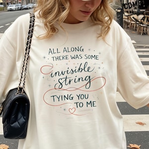 May include: Ivory-colored t-shirt with the text "ALL ALONG THERE WAS SOME invisible String TYING YOU TO ME" in a script font. The shirt is worn by a person on a crosswalk, with a black purse and a beige cap.