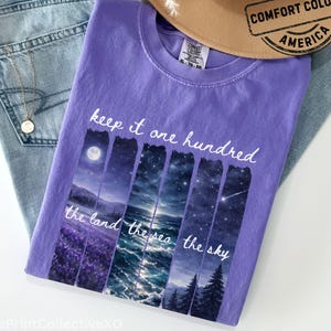 May include: A violet Comfort Colors t-shirt with the text "keep it one hundred" and "the land the sea the sky". The shirt features a landscape design with a moon, ocean, and trees. The shirt is laid on top of blue jeans and a tan hat.