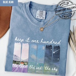May include: A blue jean Comfort Colors t-shirt with the text "keep it one hundred" and landscape art. The shirt features a necklace and a "Made in Comfort Colors America" stamp.