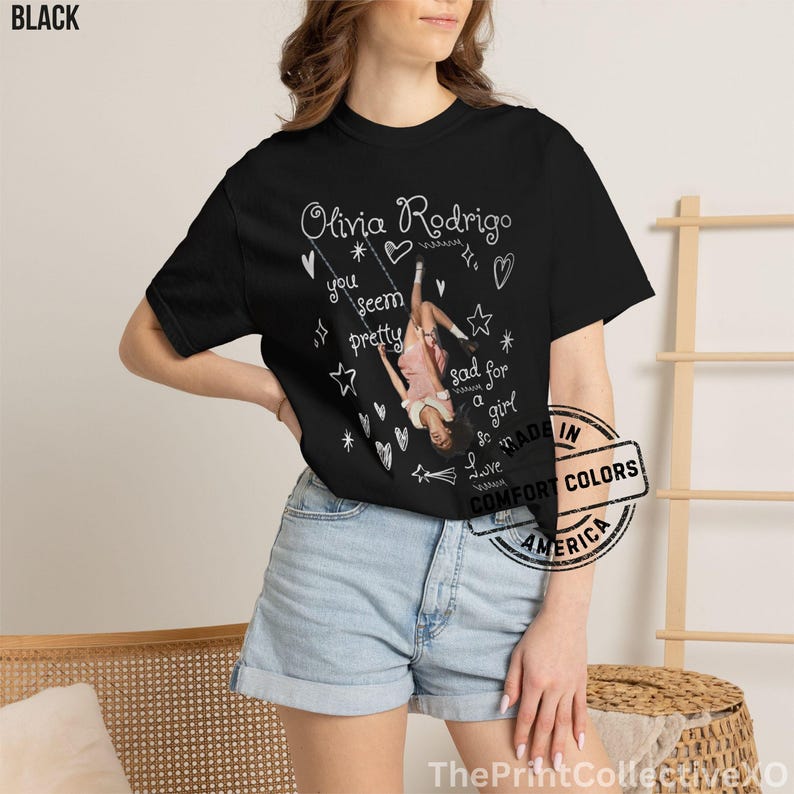 May include: Black t-shirt featuring a graphic of Olivia Rodrigo on a swing, with the text "Olivia Rodrigo" and song lyrics. The design includes hearts, stars, and the words "you seem pretty" and "sad for a girl".