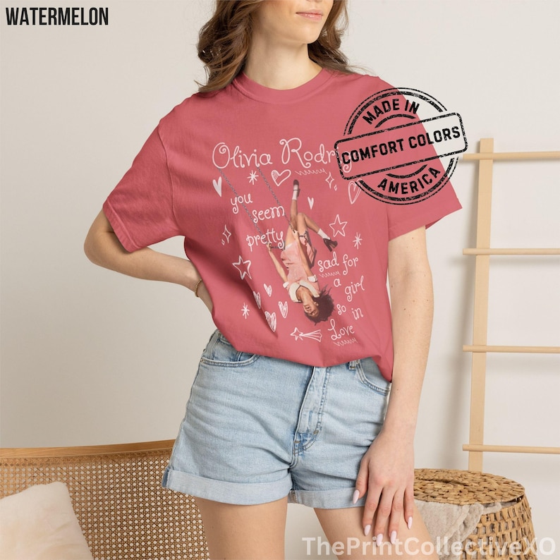 May include: Watermelon-colored t-shirt featuring a graphic of Olivia Rodrigo, with text and illustrations. The shirt has a round neckline and short sleeves. The text includes "Olivia Rodrigo" and "you seem pretty sad for a girl so in love."