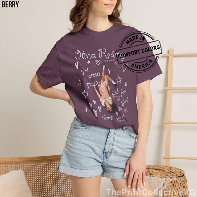 May include: Berry-colored t-shirt featuring a graphic of Olivia Rodrigo on a swing, with text that reads "you seem pretty sad for a girl so in love." The shirt also has white star and heart graphics. Made in America.