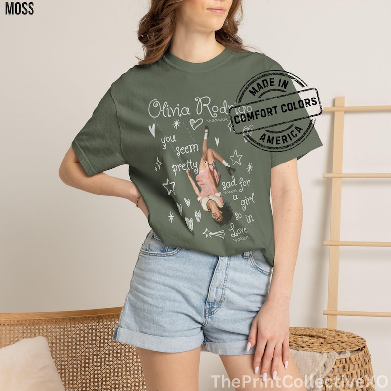 May include: Moss green t-shirt featuring a graphic of a woman in a pink dress, with the text "Olivia Rodrigo" and song lyrics. The shirt has a distressed stamp that says "Made in Comfort Colors America".