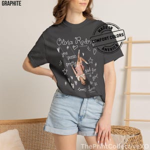 May include: Graphite-colored t-shirt featuring a graphic of Olivia Rodrigo on a swing, with the text "you seem pretty sad for a girl so in love." The shirt includes white stars, hearts, and text. Made in America.