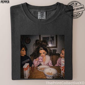May include: Pepper-colored t-shirt featuring a vintage-style image of dolls at a table. The dolls are gathered around a table with cereal and a coffee mug that says "6 AM KILL ME". The shirt has a Comfort Colors tag and a "Made in America" stamp.