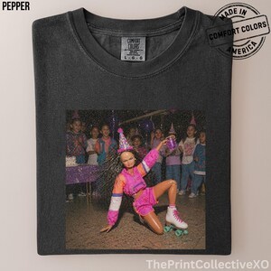 May include: Pepper-colored Comfort Colors t-shirt with a graphic print. The graphic features a doll wearing a party hat and roller skates, holding a cup, with children in the background. The shirt has a tag that says L-G-G and a stamp that says "Made in Comfort Colors America".