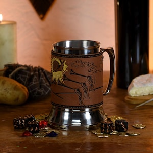 May include: A stainless steel and leather beer stein with a handle. The brown leather wrap features a sunflower and horn design. The mug sits on a wooden surface with dice, coins, and a skull.