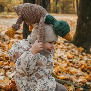 May include: A child wearing a beige jacket with a brown rabbit pattern sits on a bed of fallen leaves. The child is holding a brown and green crocheted duck plush toy above their head.
