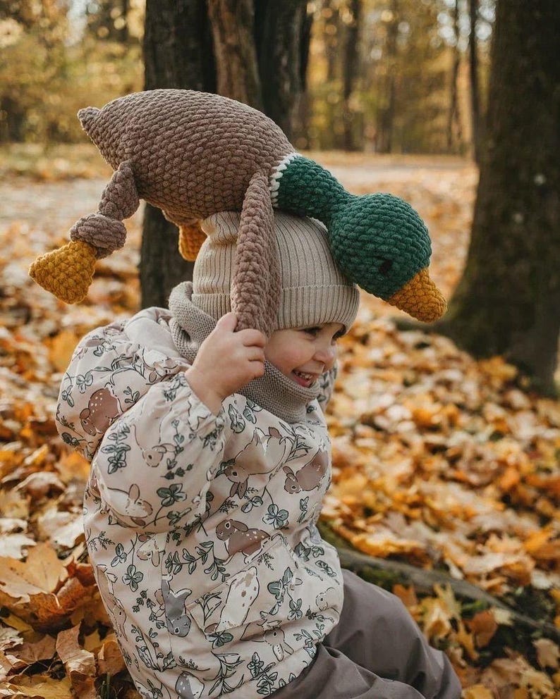 May include: A child wearing a beige jacket with a brown rabbit pattern sits on a bed of fallen leaves. The child is holding a brown and green crocheted duck plush toy above their head.