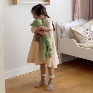 K&ouml;nnte beinhalten: Ein junges M&auml;dchen in einem hellgelben Kleid h&auml;lt ein gr&uuml;nes, geh&auml;keltes Stofftier. Sie steht auf einem Holzboden in einem Zimmer mit einem wei&szlig;en Bett im Hintergrund.