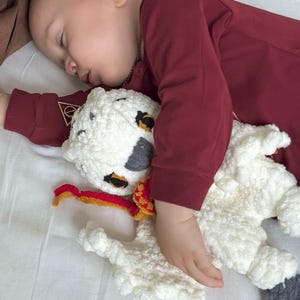 K&ouml;nnte beinhalten: Ein schlafendes Kind kuschelt mit einem handgefertigten H&auml;kellamm-Spielzeug. Das Lamm ist wei&szlig; und flauschig, mit schwarzen und grauen Flecken und einem rot-gelben Schal. Das Kind tr&auml;gt ein weinrotes Langarmshirt. Das Bild ist auf einem wei&szlig;en Laken.