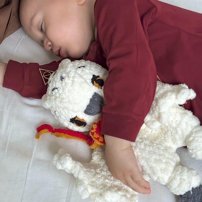 K&ouml;nnte beinhalten: Ein schlafendes Kind kuschelt mit einem handgefertigten H&auml;kellamm-Spielzeug. Das Lamm ist wei&szlig; und flauschig, mit schwarzen und grauen Flecken und einem rot-gelben Schal. Das Kind tr&auml;gt ein weinrotes Langarmshirt. Das Bild ist auf einem wei&szlig;en Laken.