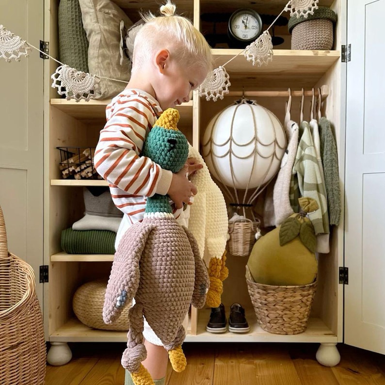 May include: A child holds a crocheted duck plush toy with brown, green, and yellow accents. The duck is standing on a wooden floor in front of a white cabinet with shelves. The cabinet is decorated with a garland of white crocheted doilies and a hot air balloon.