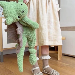 K&ouml;nnte beinhalten: Ein Kind tr&auml;gt ein hellbeiges Kleid und wei&szlig;e Sandalen und h&auml;lt ein gr&uuml;nes und braunes geh&auml;keltes Drachenspielzeug.