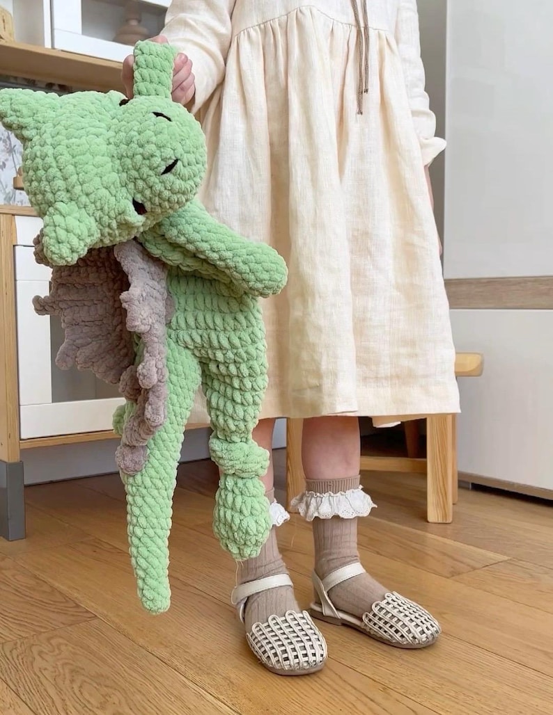 K&ouml;nnte beinhalten: Ein Kind tr&auml;gt ein hellbeiges Kleid und wei&szlig;e Sandalen und h&auml;lt ein gr&uuml;nes und braunes geh&auml;keltes Drachenspielzeug.