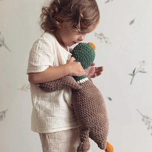 May include: A child holds a brown and green crocheted duck plush toy. The duck has orange feet and a green head with a yellow beak.