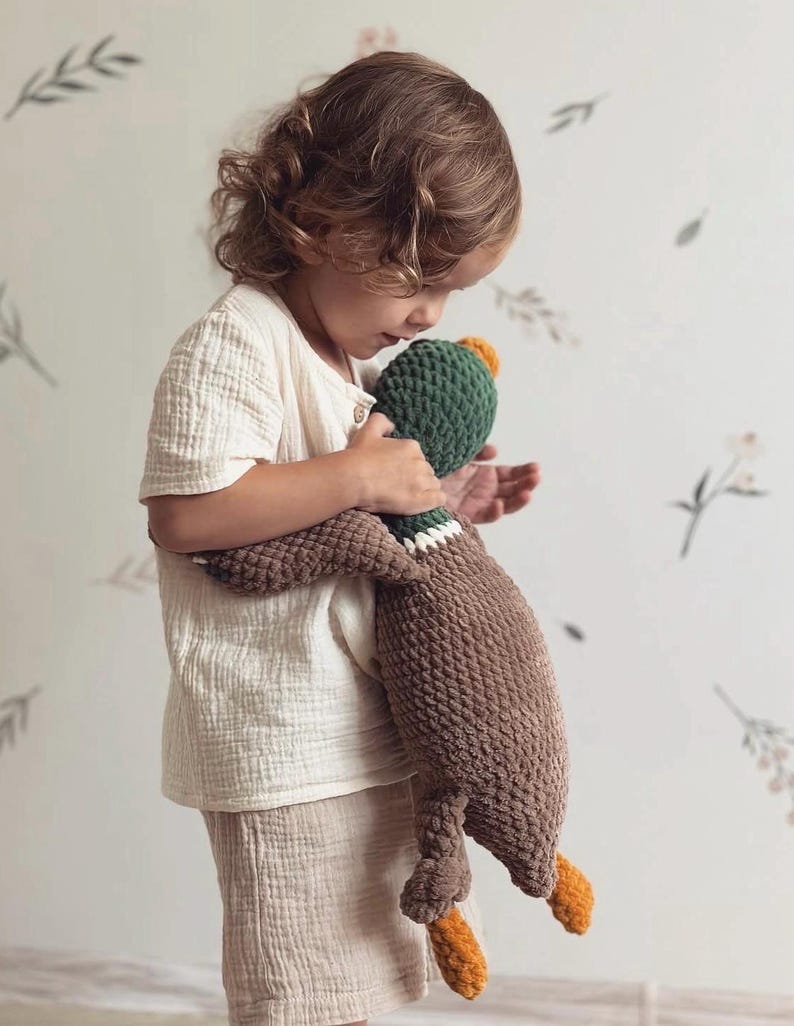 May include: A child holds a brown and green crocheted duck plush toy. The duck has orange feet and a green head with a yellow beak.