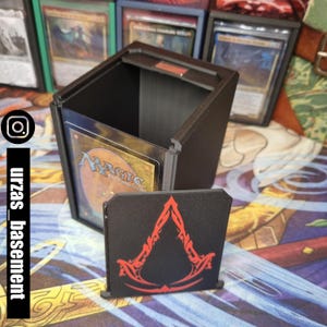 May include: A black card storage box with a clear plastic window showing a Magic: The Gathering card. The box has a black lid with a red symbol on it.