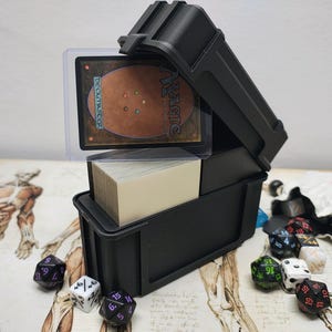 Puede incluir: Una caja de mazo de plástico negro que contiene una pila de cartas blancas, con una carta de Magic: The Gathering en una funda protectora transparente encima. Varios dados de juego poliédricos en negro, blanco y verde están esparcidos cerca.
