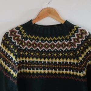 Könnte beinhalten: Ein dunkelgrüner Strickpullover mit rundem Halsausschnitt und einem mehrfarbigen Fair-Isle-Muster über Brust und Schultern. Das geometrische Design enthält horizontale Streifen in den Farben Weiß, Senfgelb und Braun.
