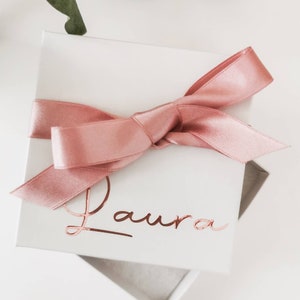 May include: A white gift box with a pink satin ribbon tied in a bow. The box is personalized with the name "Laura" in rose gold foil lettering.