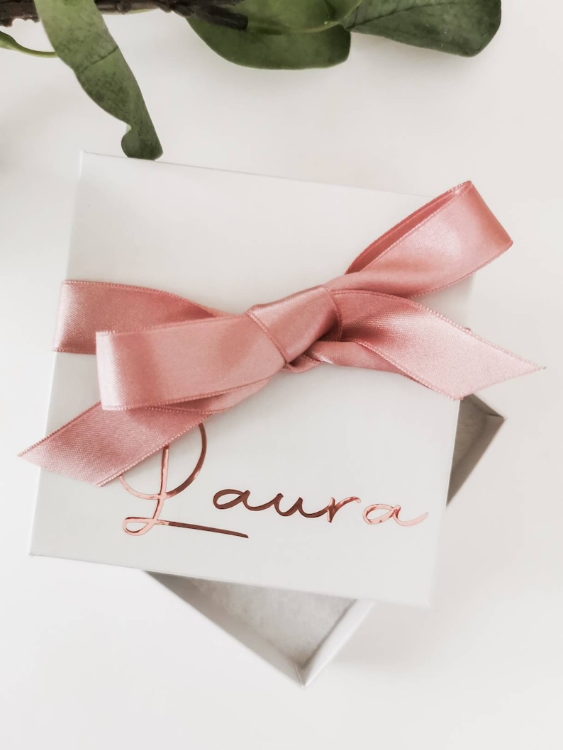 May include: A white gift box with a pink satin ribbon tied in a bow. The box is personalized with the name "Laura" in rose gold foil lettering.
