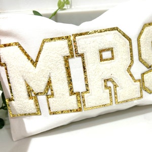 Può includere: Cuscino bianco con la parola "MRS" in lettere dorate con glitter.