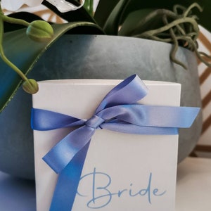 May include: A white gift box with a blue ribbon tied in a bow. The box has the word "Bride" written on it in blue lettering.