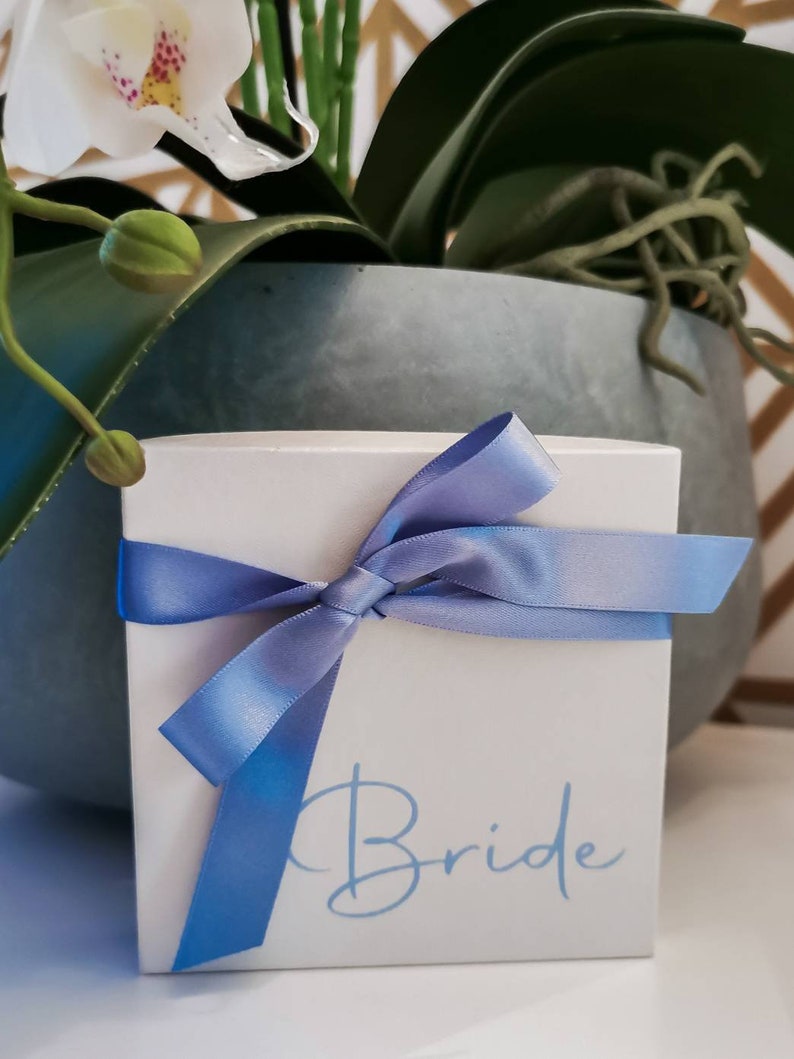 May include: A white gift box with a blue ribbon tied in a bow. The box has the word "Bride" written on it in blue lettering.