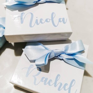 May include: Two white gift boxes with blue satin ribbon bows. The boxes are personalized with the names "Nicola" and "Rachel" in light blue lettering.