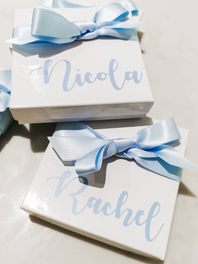 May include: Two white gift boxes with blue satin ribbon bows. The boxes are personalized with the names "Nicola" and "Rachel" in light blue lettering.