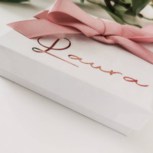 May include: A white gift box with a pink satin ribbon and bow. The box is personalized with the name "Laura" in gold foil lettering.