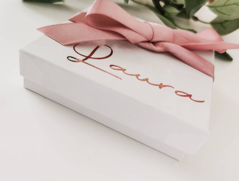 May include: A white gift box with a pink satin ribbon and bow. The box is personalized with the name "Laura" in gold foil lettering.