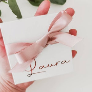 May include: A white gift box with a pink satin ribbon tied in a bow. The box is personalized with the name "Laura" in gold lettering.