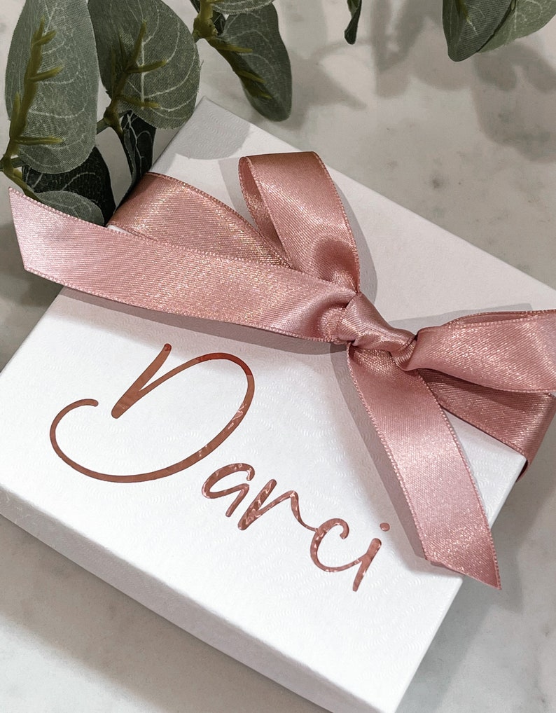May include: A white gift box with a pink satin ribbon tied in a bow. The box is decorated with the name "Danci" written in rose gold foil.