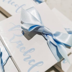 May include: Two white gift boxes with blue ribbon bows. The boxes are stacked on top of each other. The top box has the name "Rachel" written on it in blue cursive. The bottom box has the name "Nicole" written on it in blue cursive.