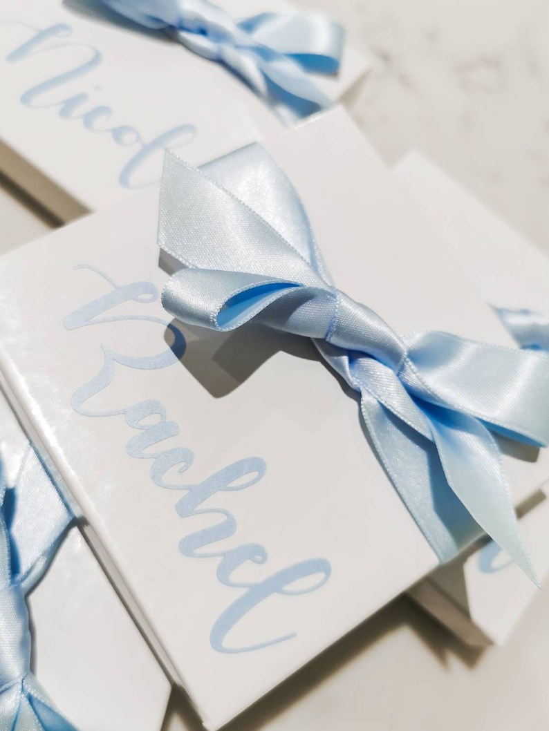 May include: Two white gift boxes with blue ribbon bows. The boxes are stacked on top of each other. The top box has the name "Rachel" written on it in blue cursive. The bottom box has the name "Nicole" written on it in blue cursive.