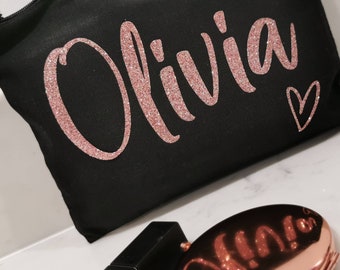 May include: Black cosmetic bag with the name "Olivia" in glittery rose gold script and a small heart. The bag is made of a textured fabric and has a zipper closure. A rose gold compact is visible in the foreground.